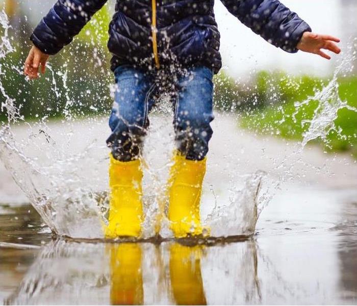 View of a child from mid chest down stomping and splashing in water puddles with yellow rainboots 